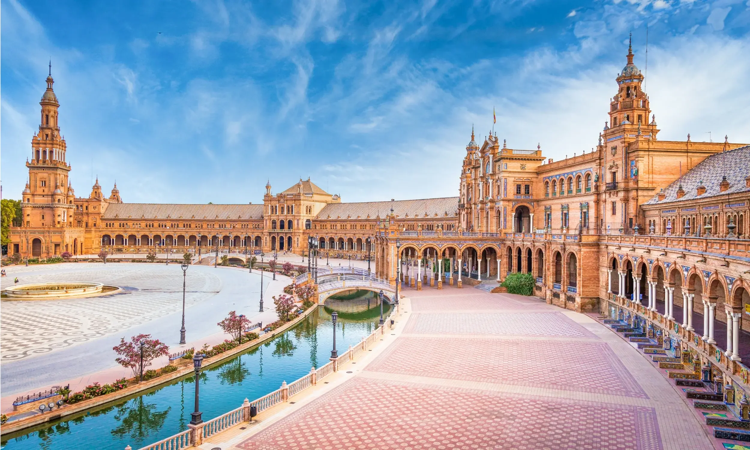 A large historic building with towers and arches, a courtyard with a fountain, a small canal, and decorative street lamps, under a blue sky with wispy clouds.