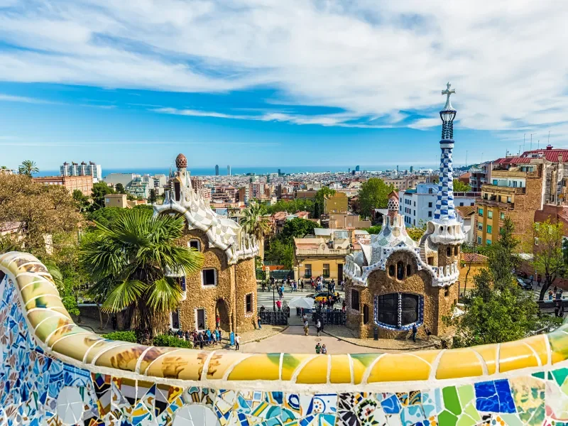 Colorful view from Park Güell in Barcelona, featuring whimsical buildings with mosaic details, a blue and white tower, lush greenery, and a city skyline with the ocean in the distance.