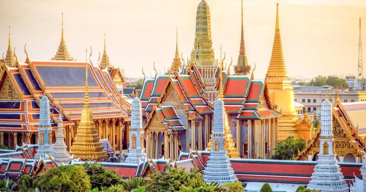 View of a Thai temple complex with multi-tiered roofs, golden spires, and ornate decorations, set against a soft sky.