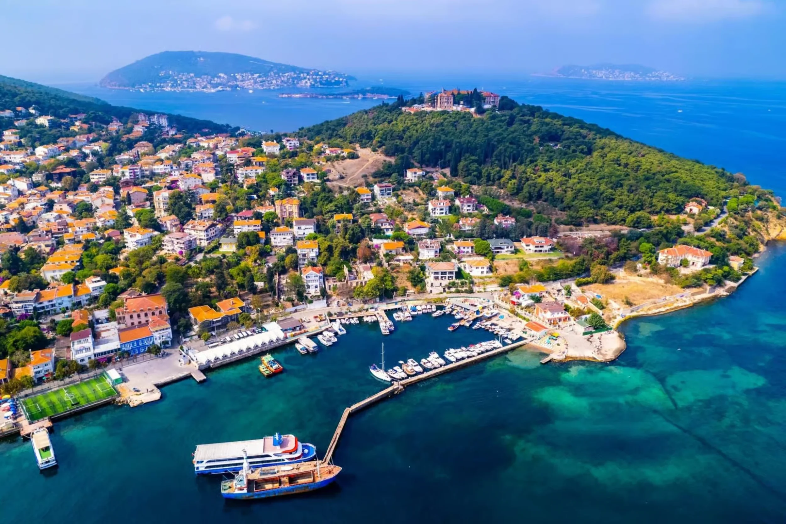 Aerial view of a coastal city with colorful houses, a marina with boats, and lush green hills extending towards the sea, with islands visible in the background.