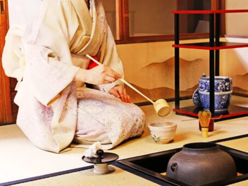 A person wearing traditional Japanese kimono, sitting on tatami mat, performing a tea ceremony with a wooden ladle, tea bowl, and ceramic pot, in a Japanese room with decorative shelves and pottery.