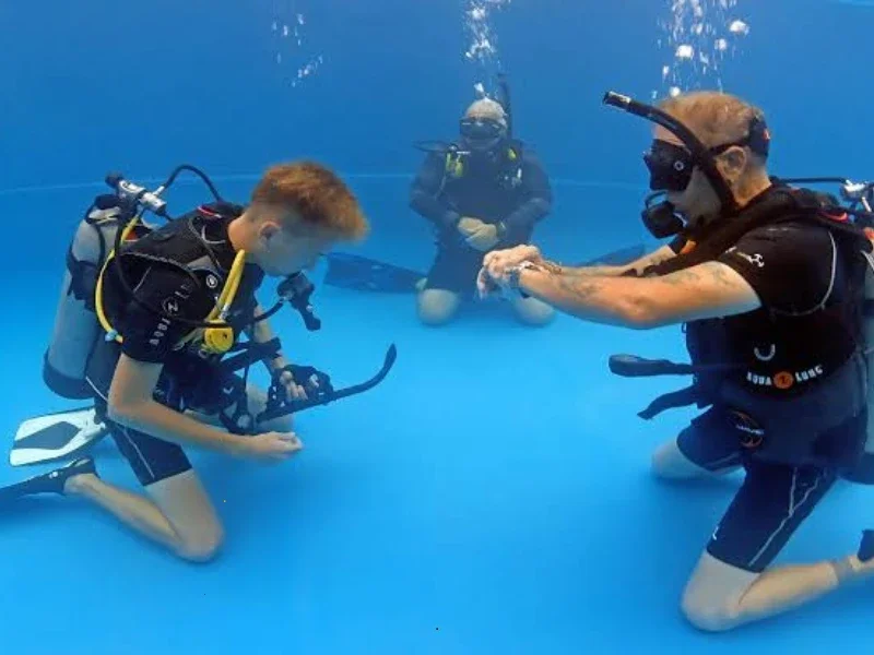 Three people, including a young boy, practicing scuba diving skills underwater in a pool with scuba tanks and masks.