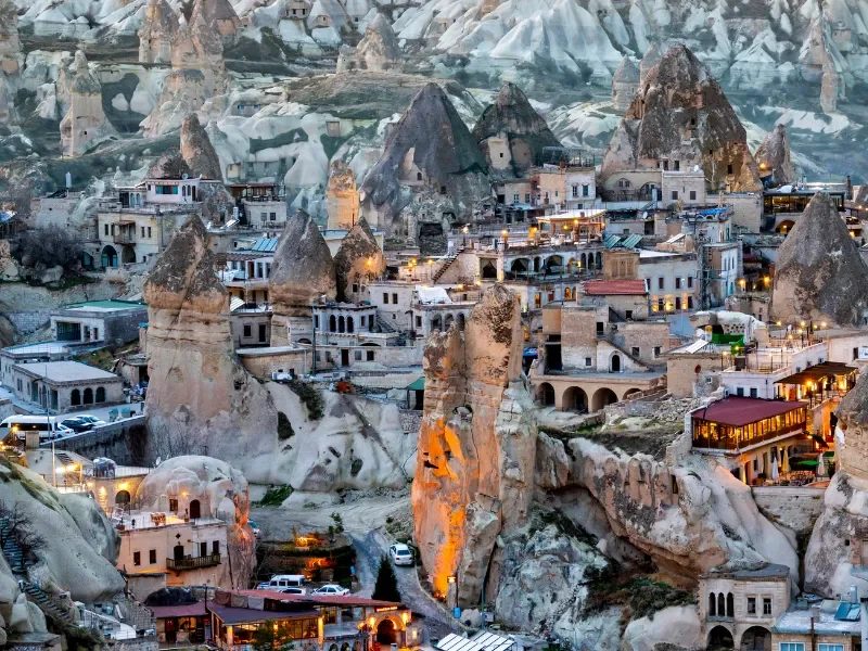 A town built into tall rock formations in Cappadocia, Turkey, with cave dwellings and illuminated pathways.