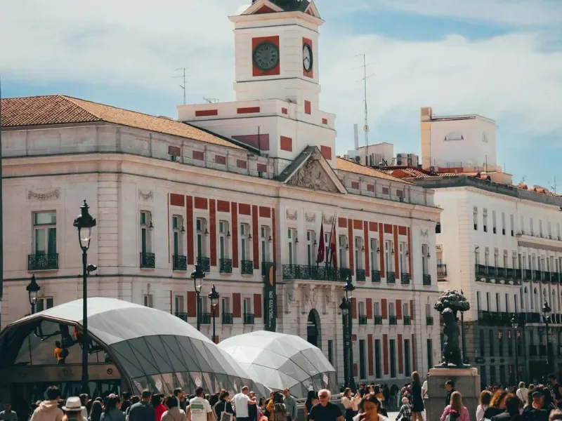 City square with historic building, clock tower, and people walking, some under umbrellas.