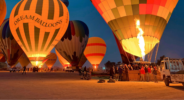 Several hot air balloons being inflated at dusk, with people and vehicles nearby. The balloons are brightly colored, with one labeled "Dream Balloons."