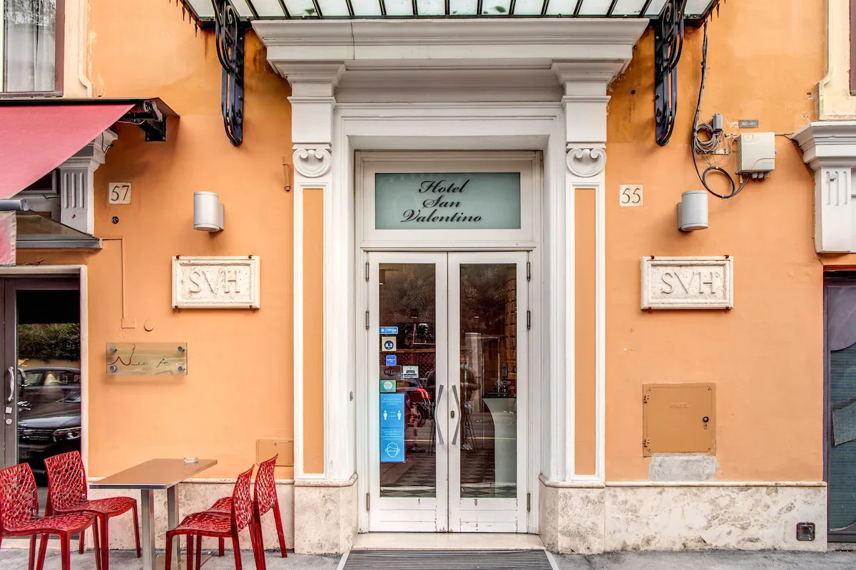 Exterior view of Hotel San Valentino with a glass door entrance, peach-colored walls, and a small outdoor seating area with red chairs and a table.