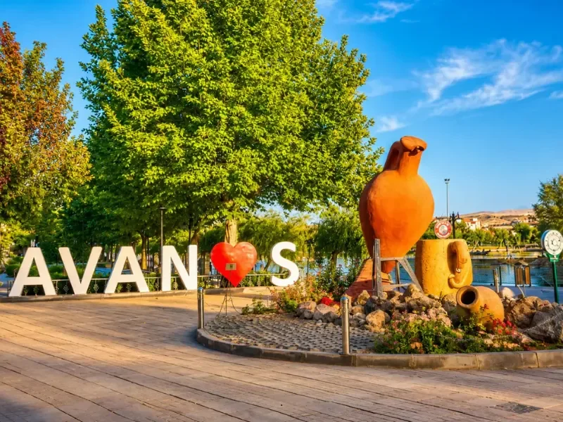 Public park with large green trees, decorative sculptures resembling large pots or vases, a white letter sign spelling 'AVAN' with a red heart in place of the letter 'A', and a lakeside view with boats and a clear blue sky.