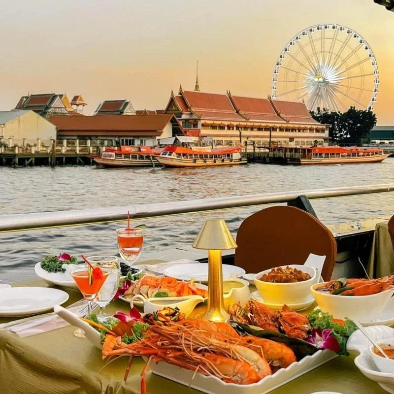 A table with seafood dishes and drinks overlooking a river with boats, traditional buildings, and a large Ferris wheel in the background during sunset.