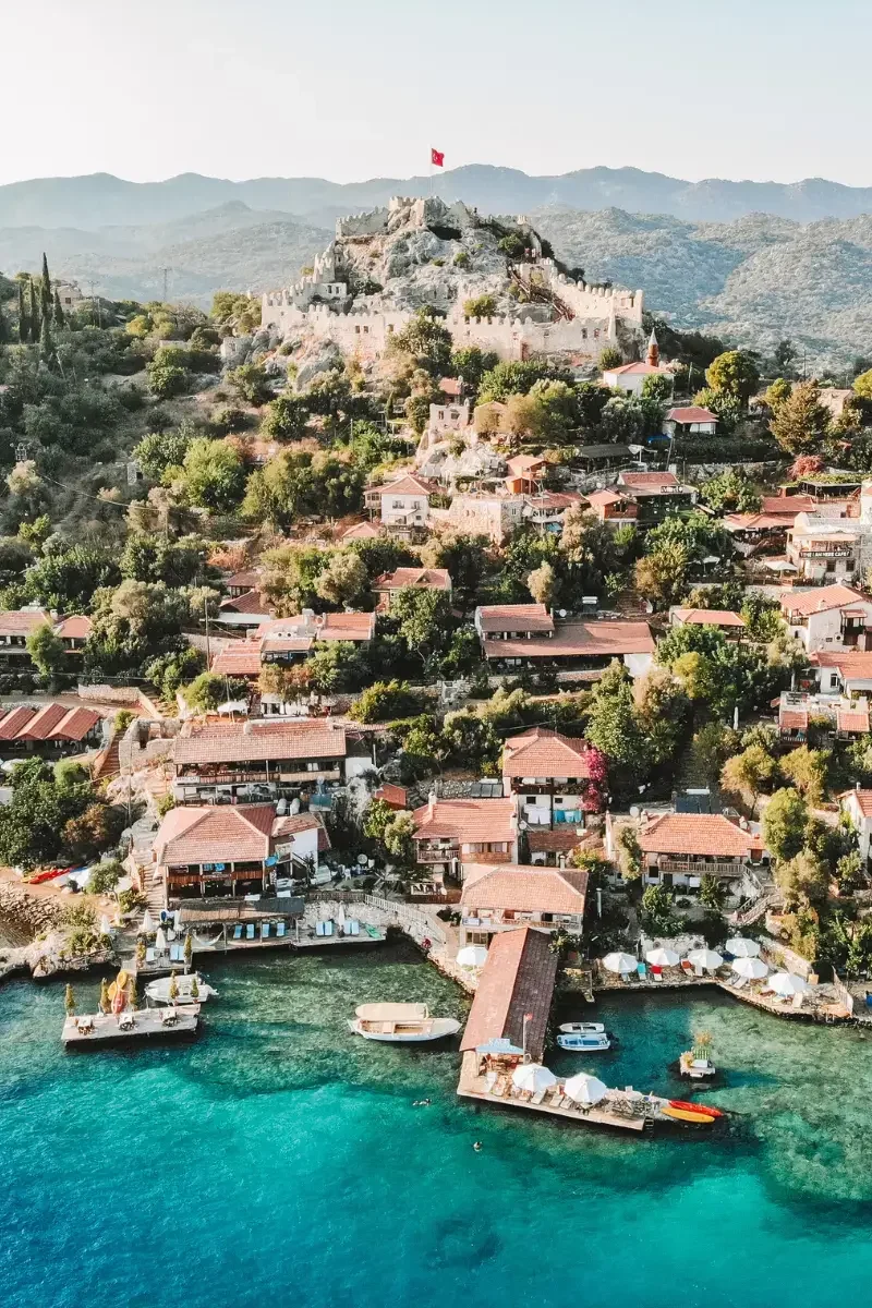 A coastal hillside town with a historic castle on top, surrounded by lush greenery and residential buildings, with a waterfront area featuring boats and umbrellas.