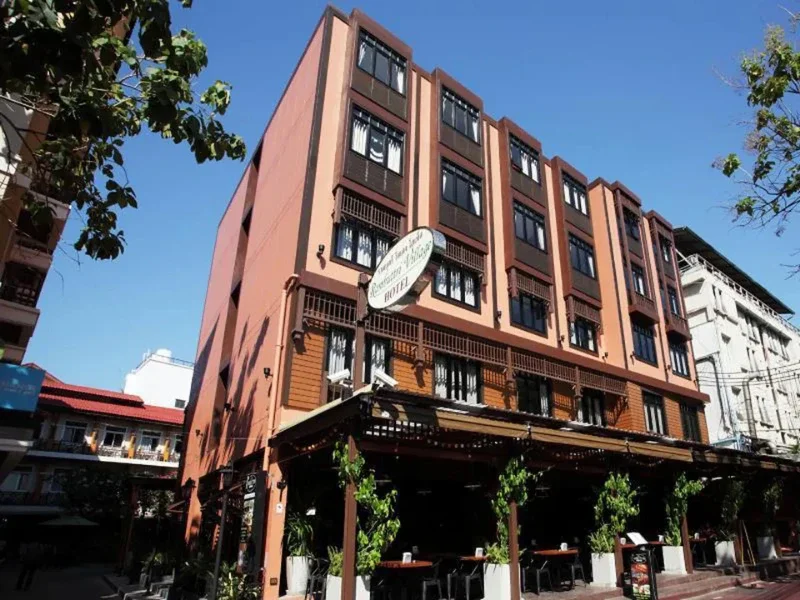 A multi-story hotel building with a reddish-brown facade, large windows, and an outdoor seating area with tables and chairs under a canopy. There are trees and other buildings nearby, and a satellite dish is visible on the hotel.