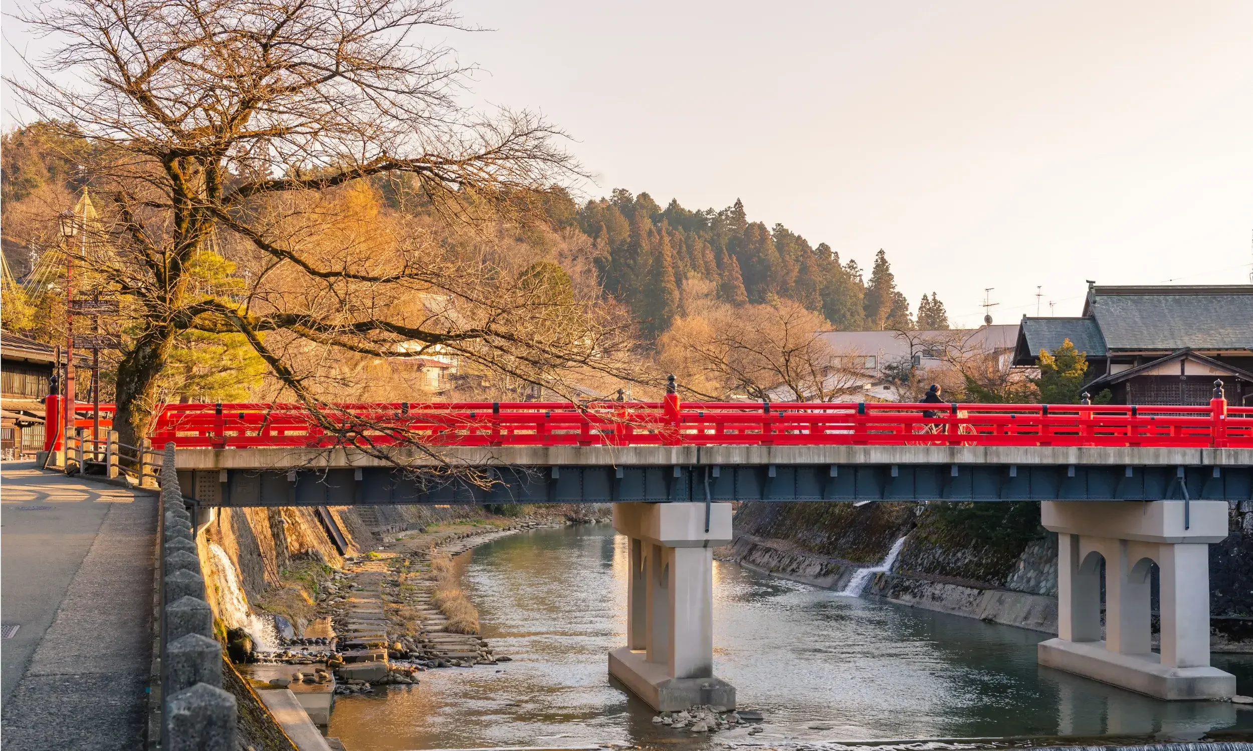 A red bridge crosses over a river in a small town, with leafless trees and houses with sloped roofs in the background, and a hillside covered with trees in the distance.