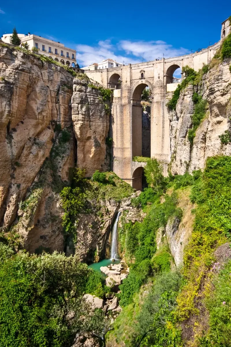 The image features a scenic view of the Puente Nuevo bridge in Ronda, Spain, spanning across a steep gorge with lush greenery at the bottom and historic buildings on top of the cliffs under a bright blue sky with some clouds.