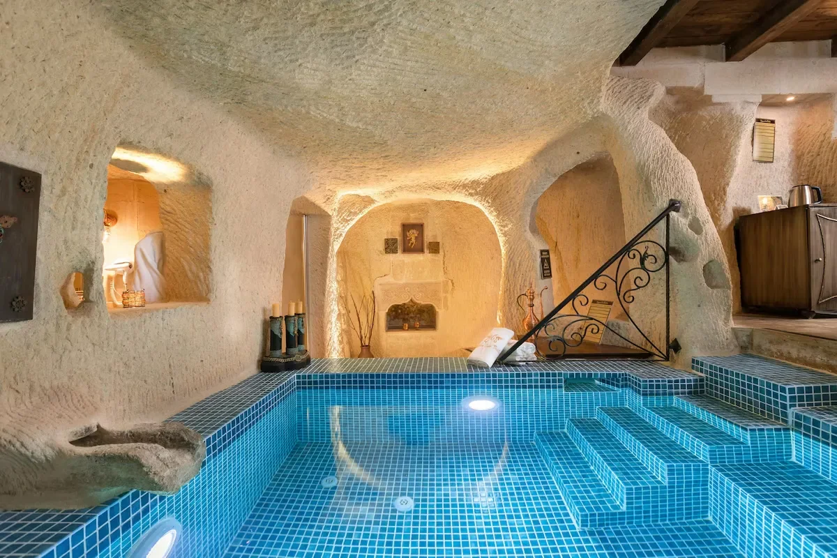 Indoor cave-style swimming pool with blue tiled steps and seating, surrounded by beige textured walls with small niches and decor, illuminated with warm ambient lighting.