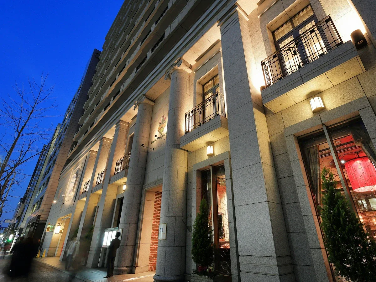 Exterior view of a multi-story building at dusk, with illuminated windows and balconies, some with black railings. Pedestrians are walking on the sidewalk, and a leafless tree is visible on the left side.