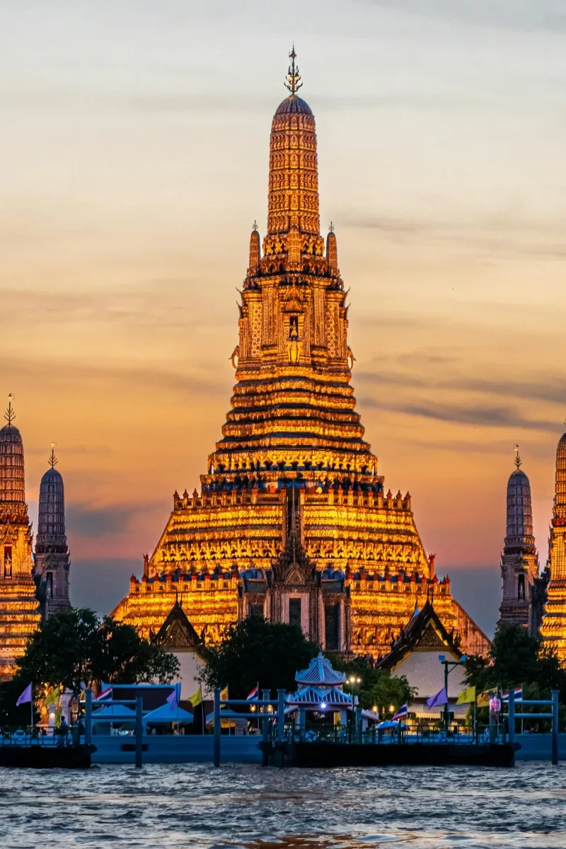 Wat Arun temple illuminated at sunset, with water in the foreground and trees at the base.