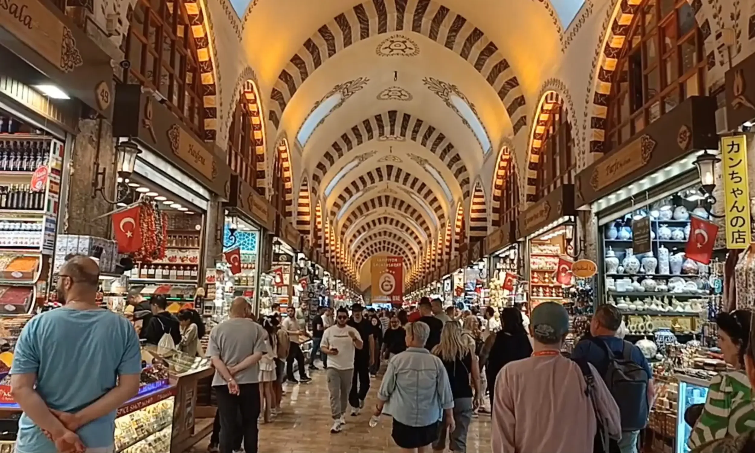Spice Bazaar, An indoor marketplace with a high, arched ceiling decorated with intricate patterns, bustling with shoppers and vendors. Shops display various goods including pottery, souvenirs, and snacks, with Turkish flags hanging around.