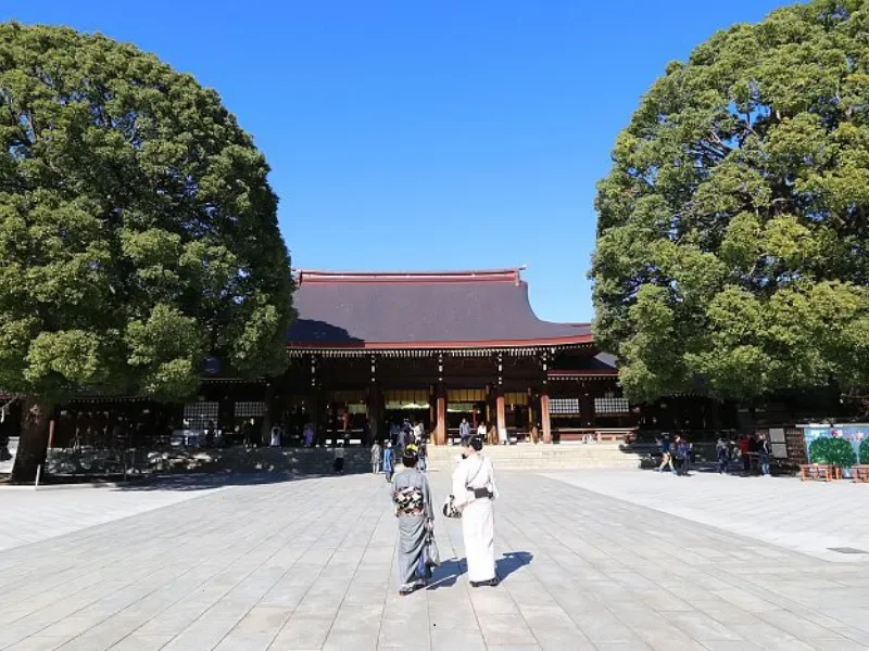 Traditional Japanese temple with wooden architecture, surrounded by green trees, under a clear blue sky, with visitors walking in the courtyard.