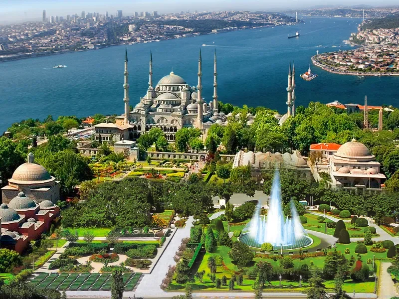 Aerial view of a large mosque with multiple minarets, a water fountain, lush gardens, and surrounding buildings, with a river and city skyline in the background.