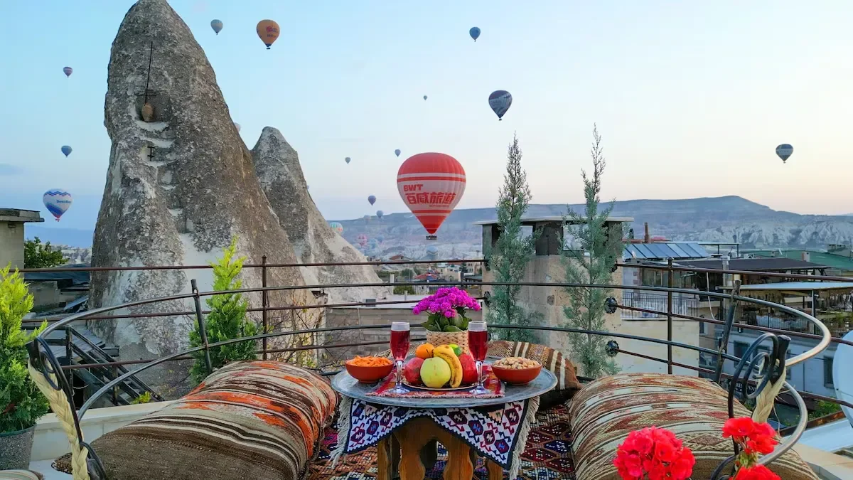 Balcony with a table set with a flowerpot, fruits, and drinks, overlooking a landscape with rock formations and hot air balloons in the sky.