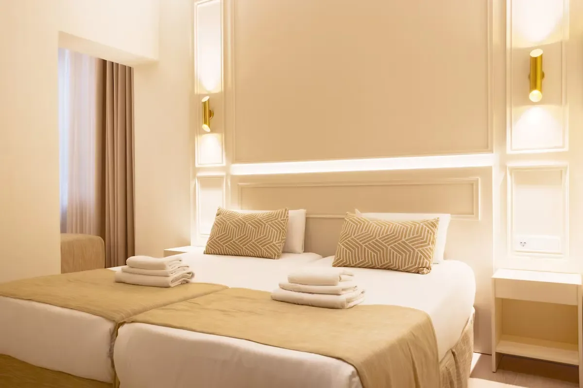 A neatly made hotel bed with two pillows, folded towels, a beige bed runner, and a bedside table. The room has soft lighting and beige and white decor.
