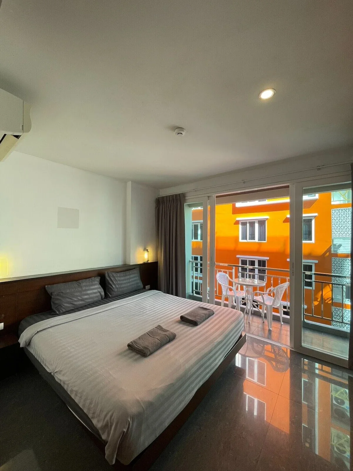 A hotel room with a large bed, dark wood headboard, gray pillows and bedsheets, and two towels placed on the bed. There is a small balcony with outdoor furniture, including a round table and four chairs. The room has large sliding glass doors leading to the balcony, and an orange building with windows is visible outside.