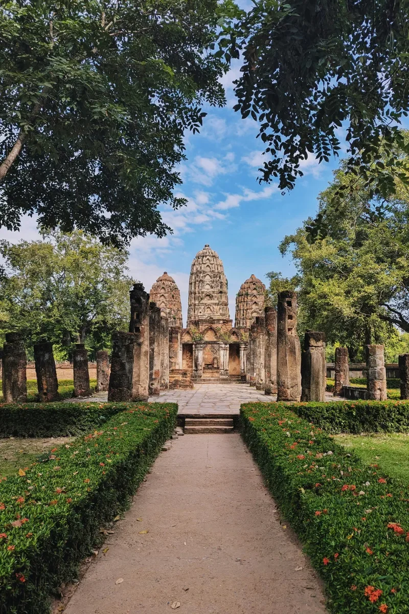 Ancient temple ruins with three tall spires, surrounded by columns and lush greenery, under a blue sky with some clouds.