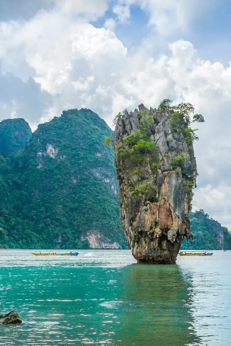 A tall, rocky limestone island with green vegetation, rising from turquoise waters with boats passing nearby, set against lush mountains and a cloudy sky.