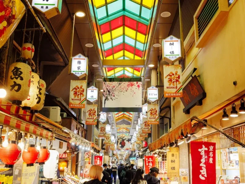 Indoor shopping street with colorful stained glass ceiling, lanterns, and signs in Japanese, busy with shoppers.