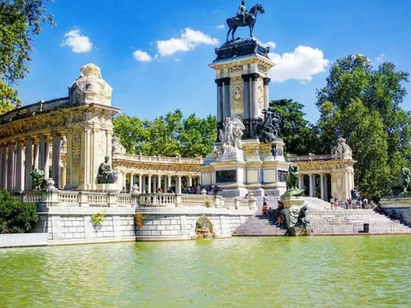 A grand ornate monument with sculptures, a large central column, and an equestrian statue at the top, surrounded by water and green trees under a blue sky.