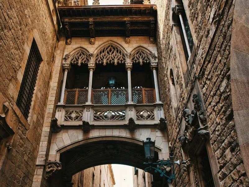 An ornate Gothic-style balcony with intricate wood carvings and arched windows, situated between old stone buildings with totem sculptures below.
