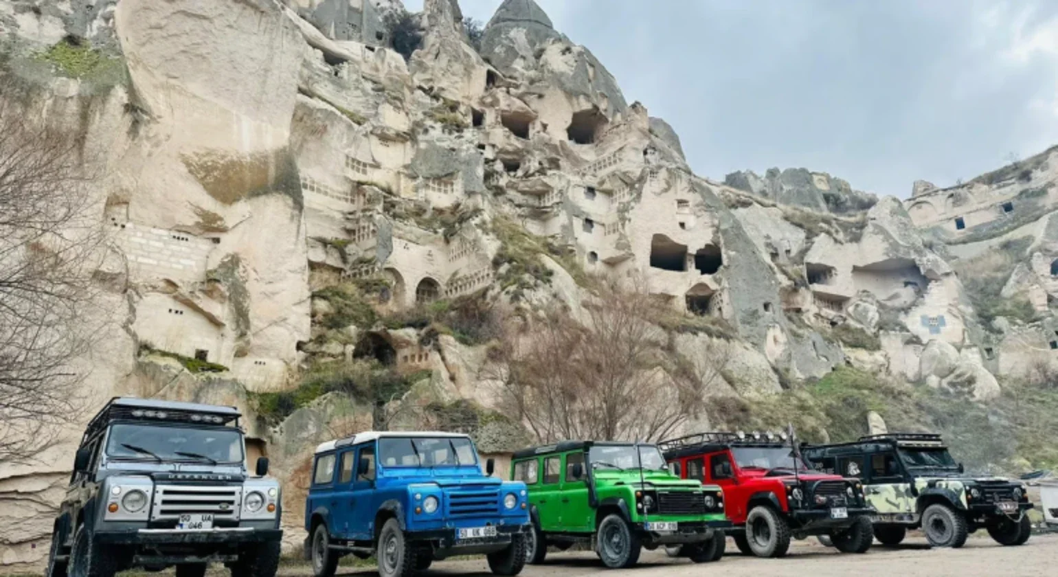 Five off-road vehicles parked in front of a hill with numerous caves and carved openings in the rock.
