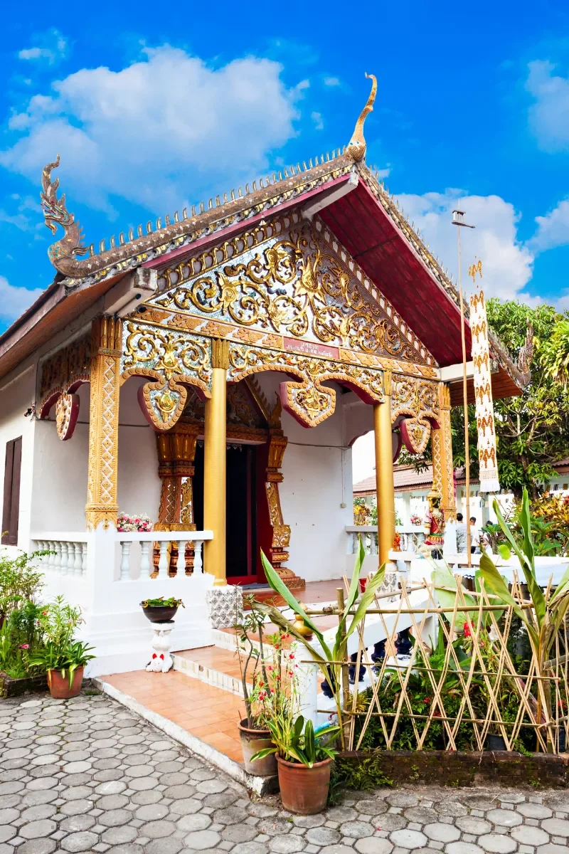 A traditional Thai temple with ornate gold and red decorations on the roof and columns, surrounded by potted plants and a blue sky with clouds in the background.