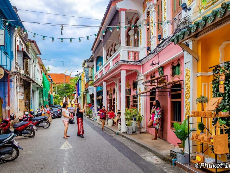 Colorful street scene with pastel-colored buildings, people walking and socializing, motorcycles parked along the road, and decorative lights hanging overhead.