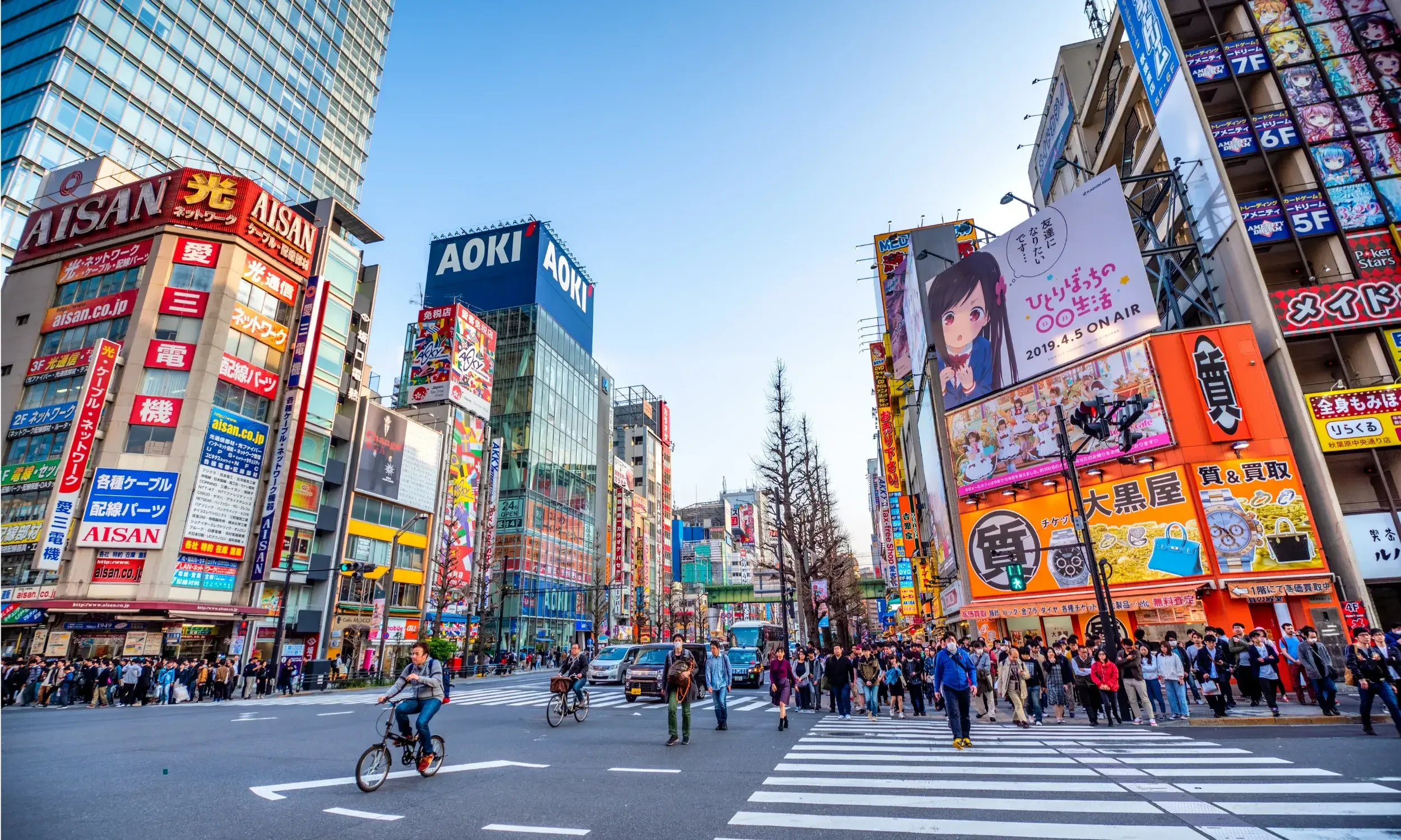 A busy city street in Japan with numerous colorful billboards and signs on buildings. People are crossing the street, and cyclists are riding through the intersection. The sky is clear and blue, and the scene is vibrant with advertisements and tall buildings lining both sides of the street.
