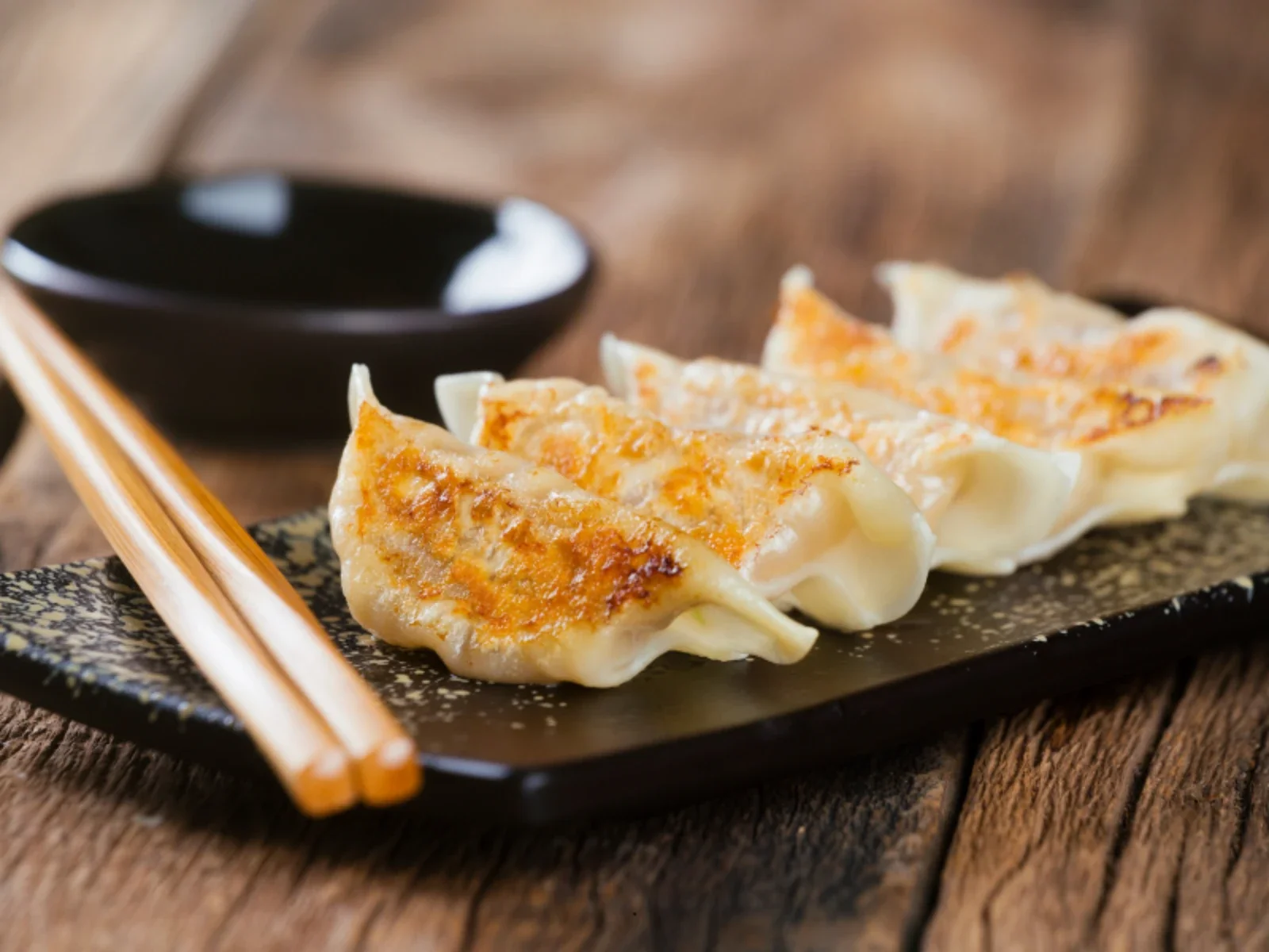 Close-up of three pan-fried dumplings on a black rectangular plate with a textured surface, a small black bowl of soy sauce, and wooden chopsticks on a rustic wooden table.