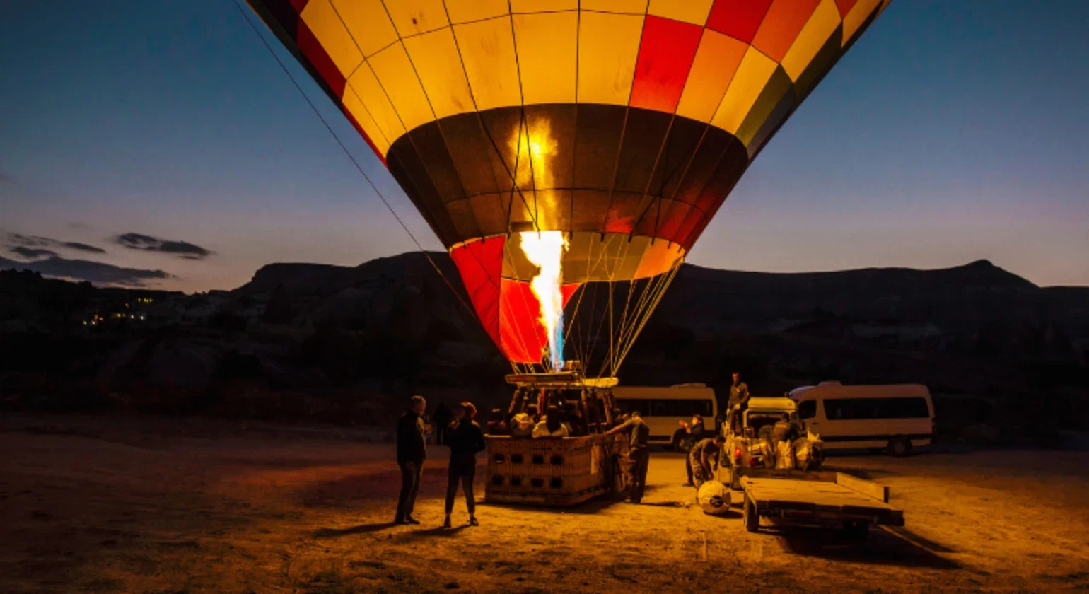 A colorful hot air balloon being inflated at dusk with a flame from the burner illuminating the basket and fabric. Several people are gathered around, preparing the balloon for flight in an open field with hills in the background.