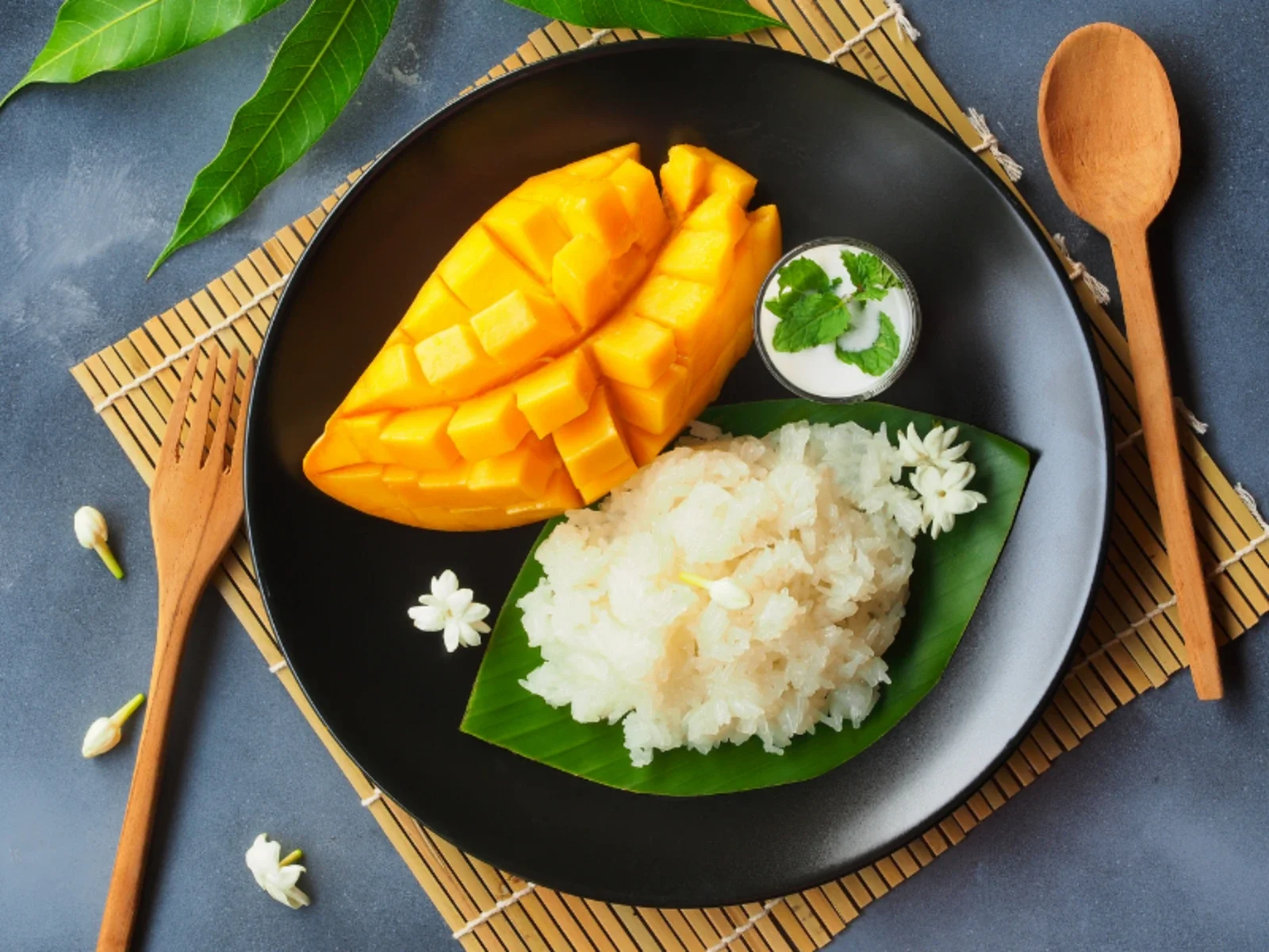 A plate of Thai mango sticky rice with sliced mango, sticky rice, coconut milk, and garnishes. The plate is on a black surface with wooden utensils and green leaves.