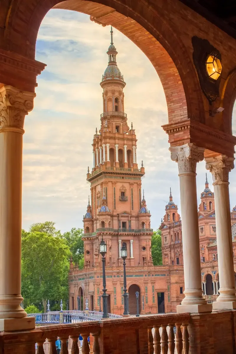 A historic pink-colored tower with ornate architecture and multiple spires, seen through an arched balcony with classical columns and decorative railing, surrounded by trees and street lamps.