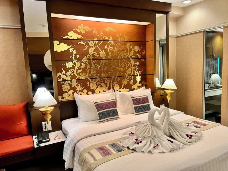 A hotel room with a neatly made bed, two swan towels decorated with purple flowers, decorative pillows, a wooden headboard with gold floral design, bedside lamps, and a mirror on the wall.