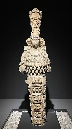 Ancient stone sculpture of a humanoid figure with elaborate headdress and detailed ornamentation, displayed in a museum setting.