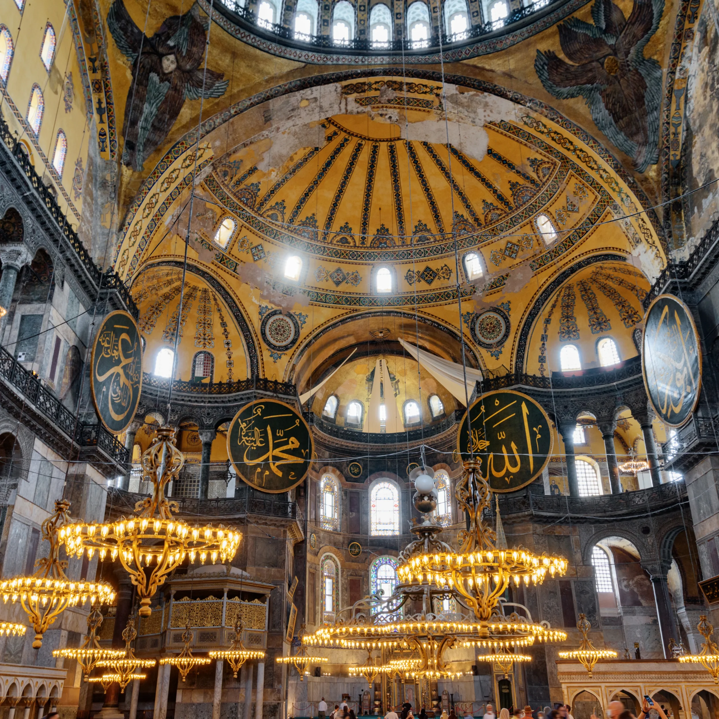 Interior of a historical mosque showcasing a large, ornate gold and blue ceiling with intricate patterns, chandeliers hanging from it, tall windows, and visitors walking inside.