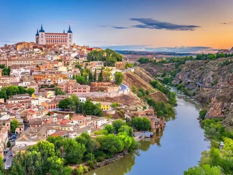 A sprawling city built on a hillside with a river running through a canyon, featuring historic buildings, greenery, and a prominent castle-like structure at the top of the hill during sunset.