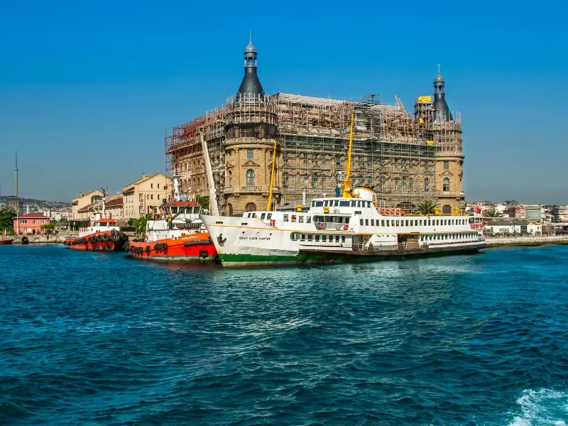 View of a historic building under renovation with scaffolding, situated along a waterfront with boats docked in front, under a clear blue sky.