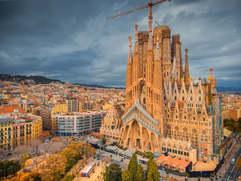Aerial view of the Sagrada Família, a large unfinished Roman Catholic basilica in Barcelona, Spain, under construction with cranes, surrounded by city buildings and a cloudy sky.