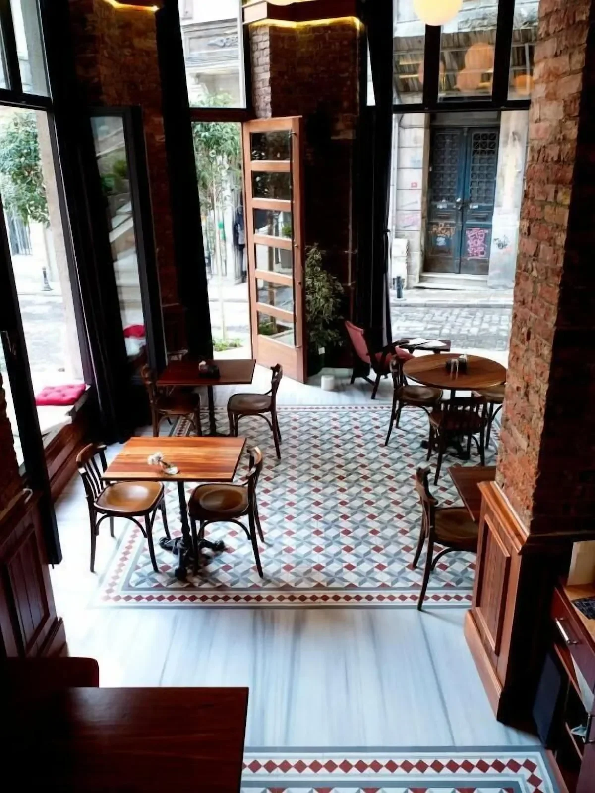 Interior view of a cozy cafe with natural light coming through large windows, wooden tables and chairs, patterned tile flooring, and brick walls. Open door shows a cobblestone street outside.