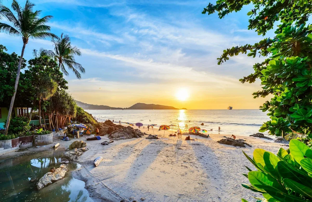 A tropical beach at sunset with palm trees, colorful umbrellas, and people enjoying the water and sand.