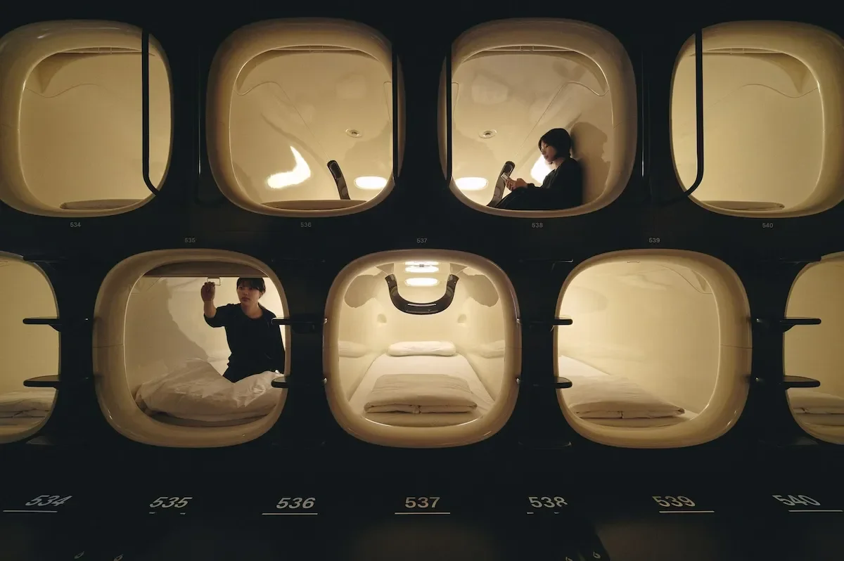 Multiple capsule sleeping pods arranged in a grid, with two women inside, one on her phone and the other making a signing gesture.