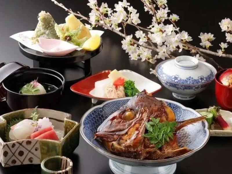 Traditional Japanese meal with various dishes including a cooked fish, sashimi, tempura, rice, pickles, and miso soup, arranged on a dark table with cherry blossoms in the background.