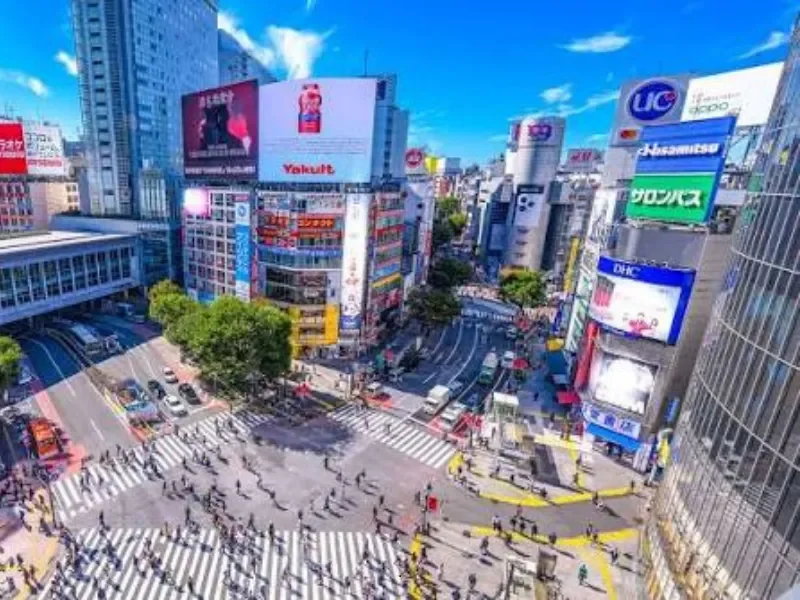 A busy city intersection with pedestrians crossing multiple crosswalks, surrounded by tall buildings with digital billboards and advertisements, under a partly cloudy blue sky.