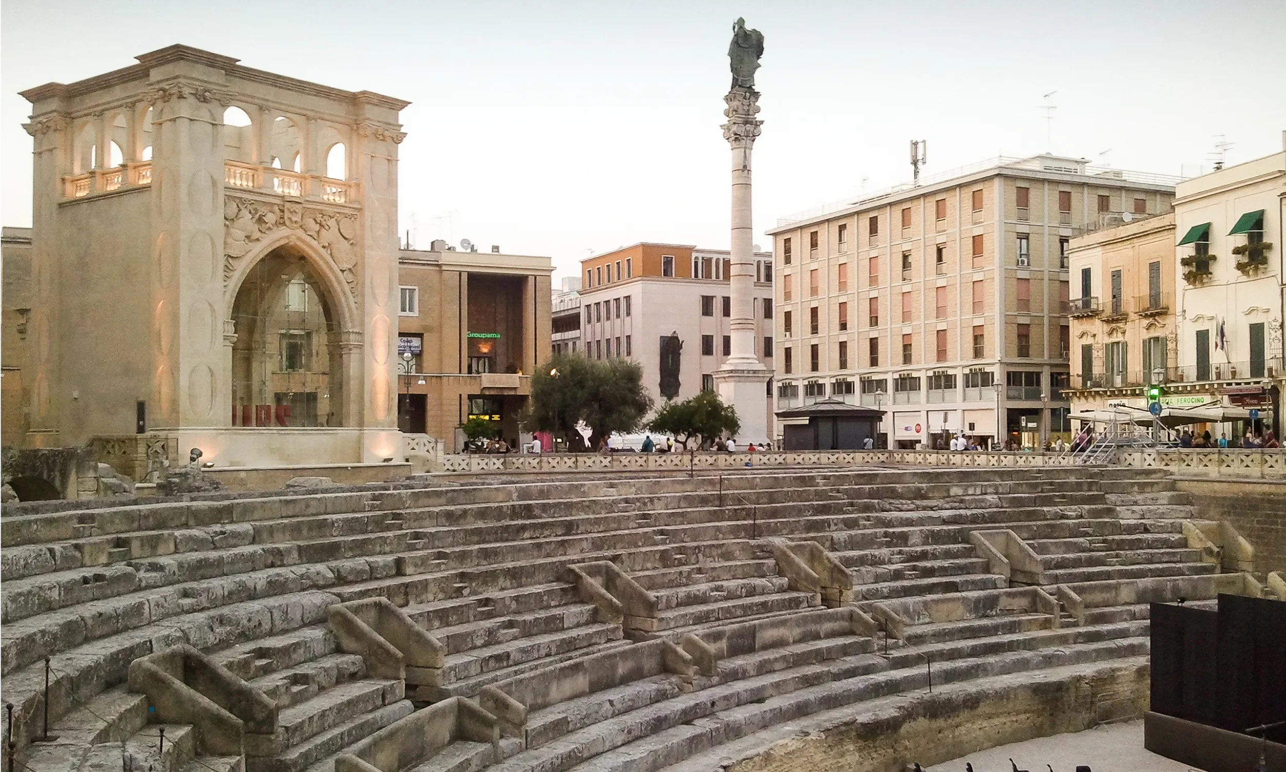 An archaeological site with ancient stone steps leading up to a plaza, featuring a large stone arch, a tall column with a statue on top, and modern buildings in the background, including a bank and shops.
